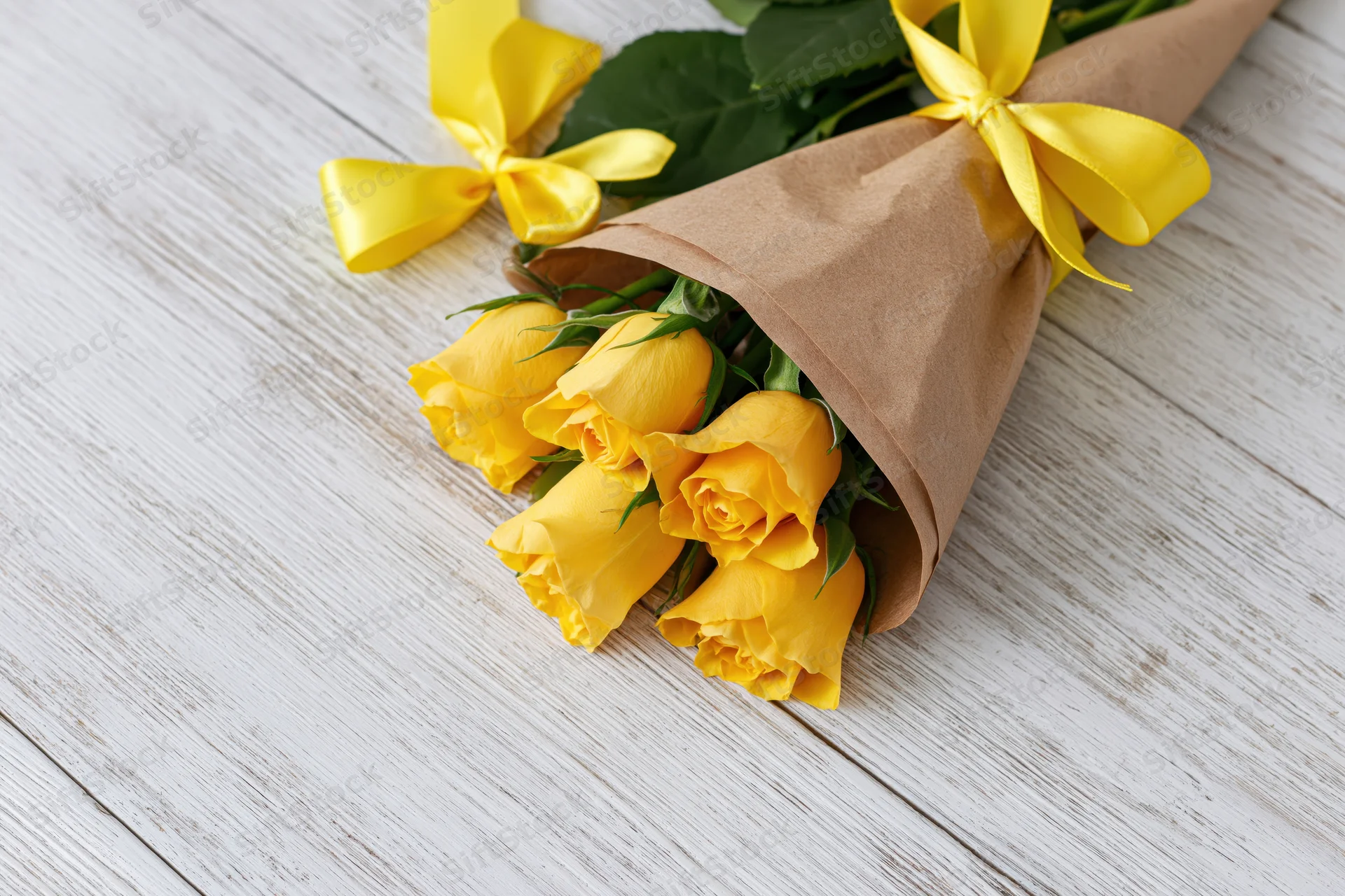 Small bouquet of yellow roses wrapped in brown paper cone with yellow ribbon on wooden surface