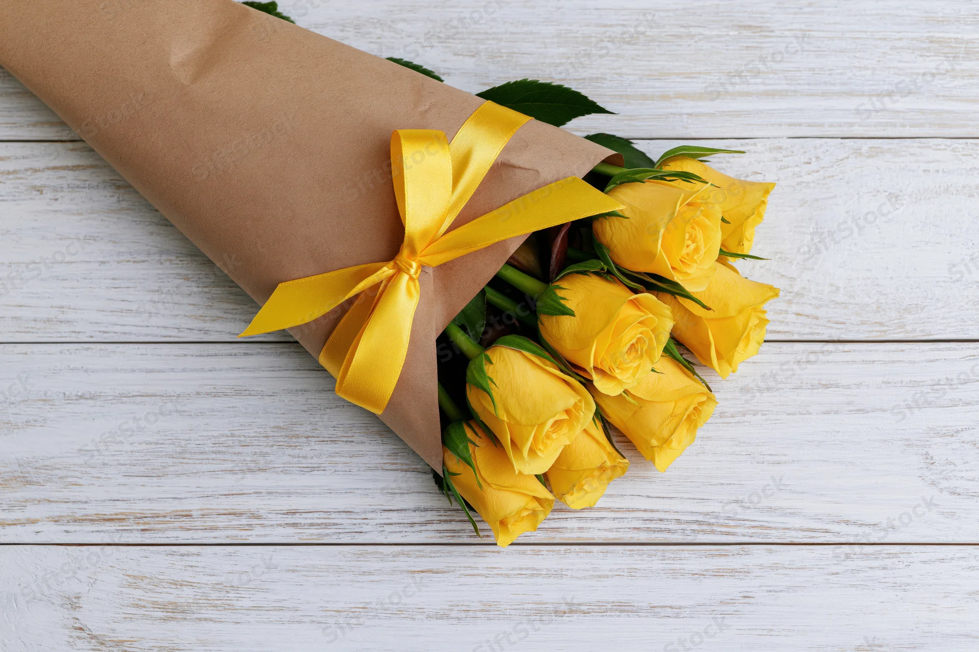 Small bouquet of yellow roses wrapped in brown paper cone with yellow ribbon on white wooden surface