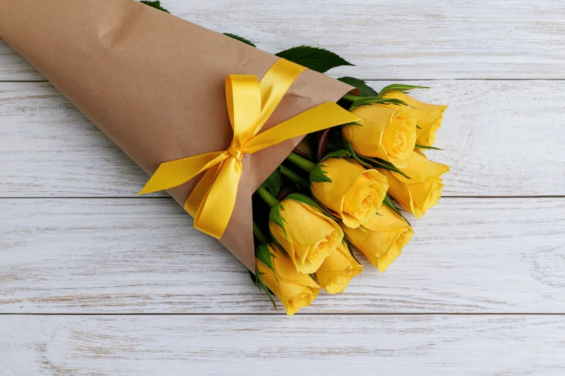 Small bouquet of yellow roses wrapped in brown paper cone with yellow ribbon on white wooden surface