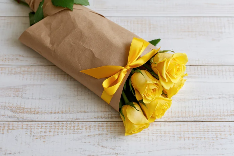 Small bouquet of yellow roses wrapped in brown paper with yellow ribbon on white wooden surface