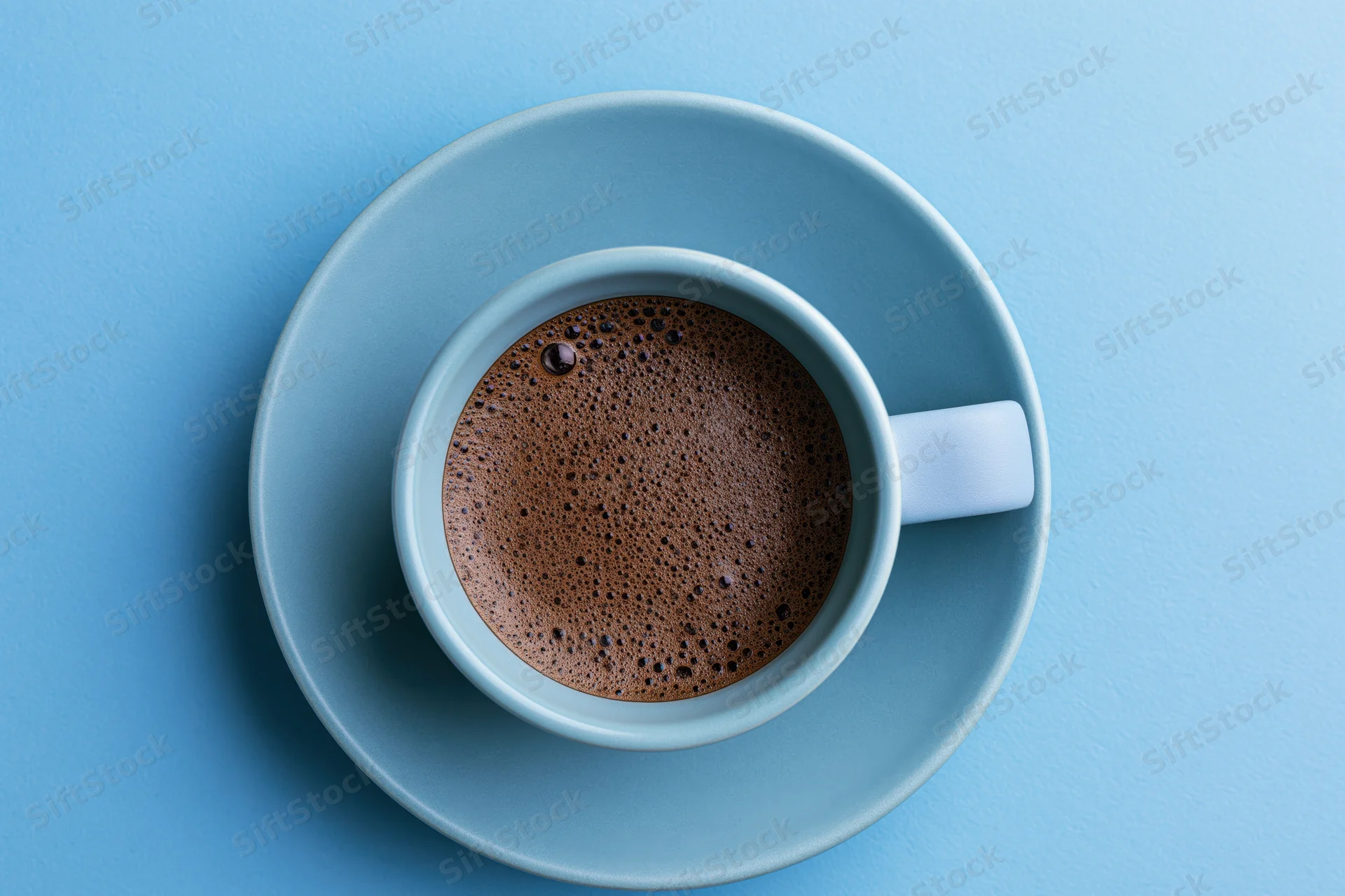 Light-blue ceramic cup filled with black espresso coffee on matching saucer, viewed from above on solid blue background