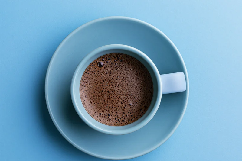 Light-blue ceramic cup filled with black espresso coffee on matching saucer, viewed from above on solid blue background