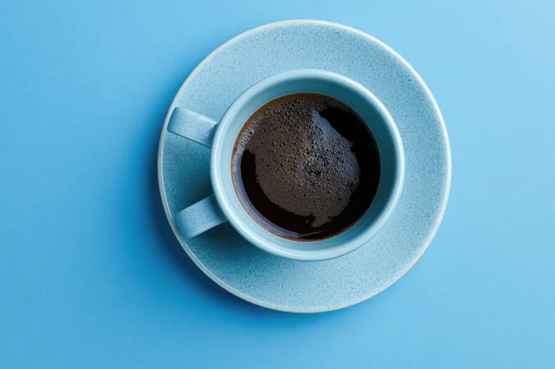 Small light-blue ceramic cup filled with black espresso on matching saucer against solid blue background, top-down view
