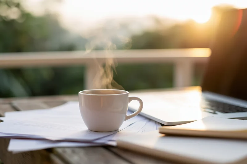 Steaming white ceramic coffee cup on scattered papers beside laptop on wooden balcony table at sunrise