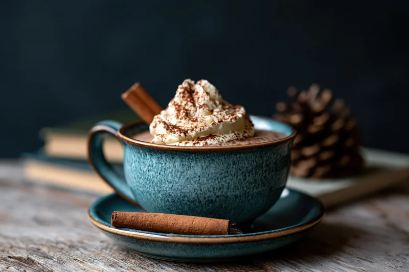 Teal ceramic mug filled with hot chocolate topped with whipped cream and cinnamon, on a wooden table with a pinecone and cinnamon stick nearby