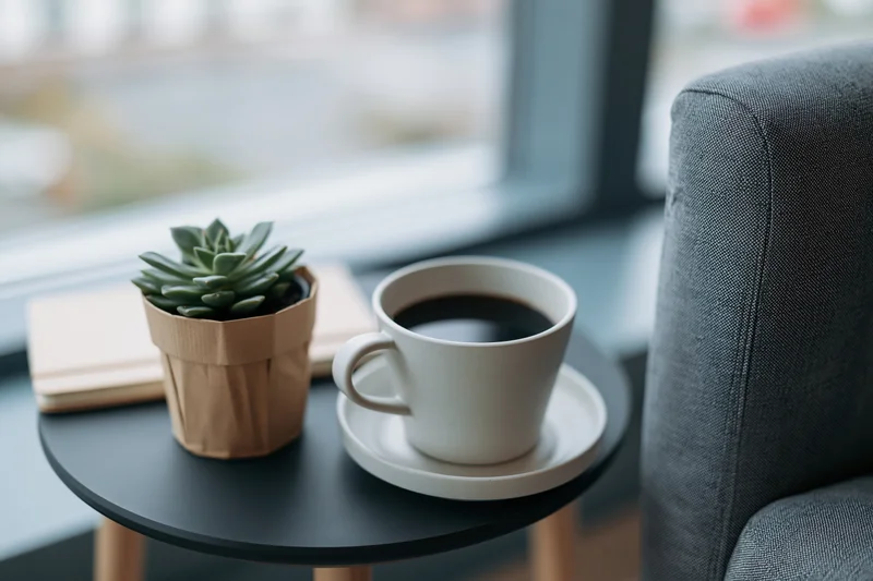 White ceramic coffee cup with black coffee on a small round table beside a potted succulent and notebook near a window