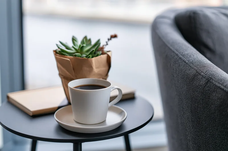 White ceramic coffee cup with black coffee on saucer on small round table beside succulent plant and notebook near window