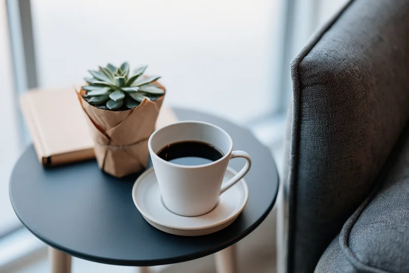 White ceramic coffee cup with black coffee on a small round table beside a gray sofa, with a succulent plant and notebook nearby