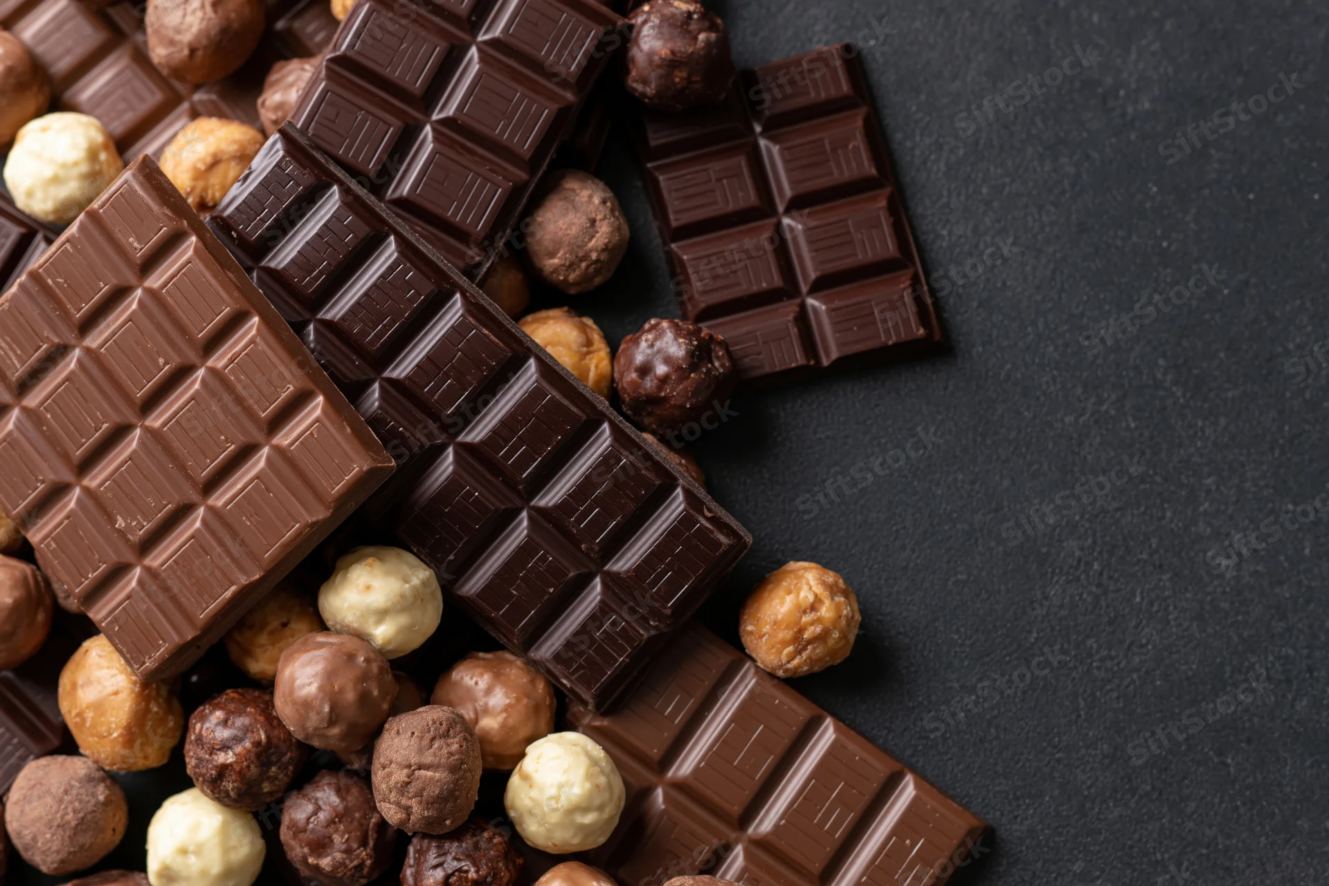 Assorted chocolate bars and pralines arranged in a loose pile on a dark surface with copyspace
