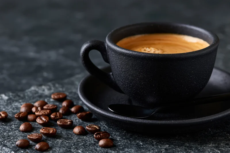 Black ceramic coffee cup filled with espresso crema on saucer with scattered beans on stone surface