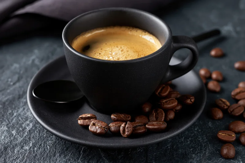 Black ceramic coffee cup filled with espresso crema on saucer with scattered coffee beans on dark stone surface