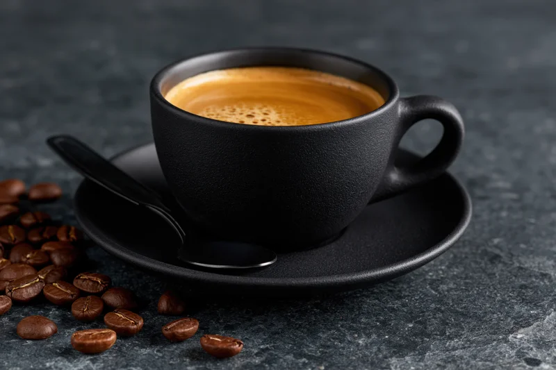Black ceramic coffee cup filled with espresso crema on saucer with spoon and scattered beans on dark stone surface