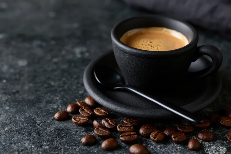 Black ceramic coffee cup with espresso crema and scattered beans on dark textured surface