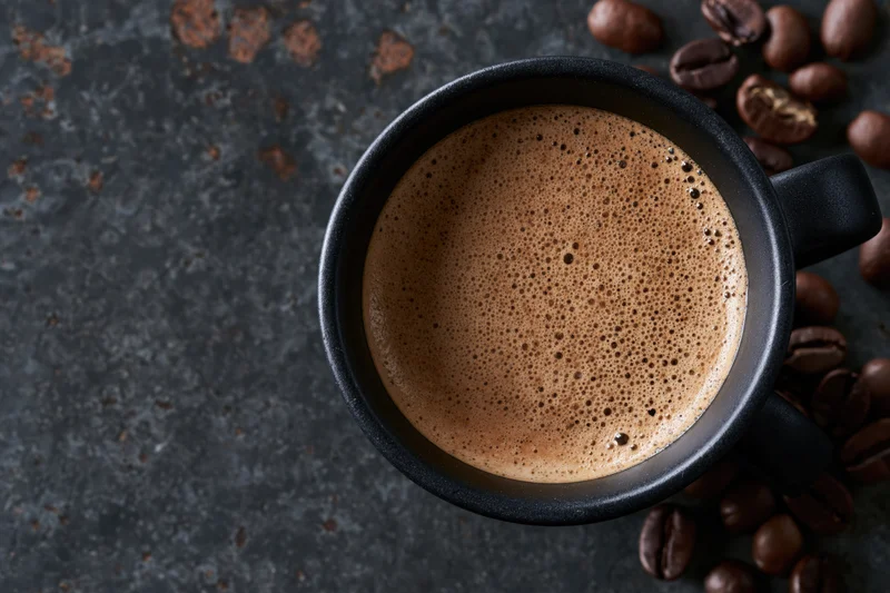 Black ceramic mug filled with frothy espresso coffee viewed from above on dark textured surface with scattered coffee beans