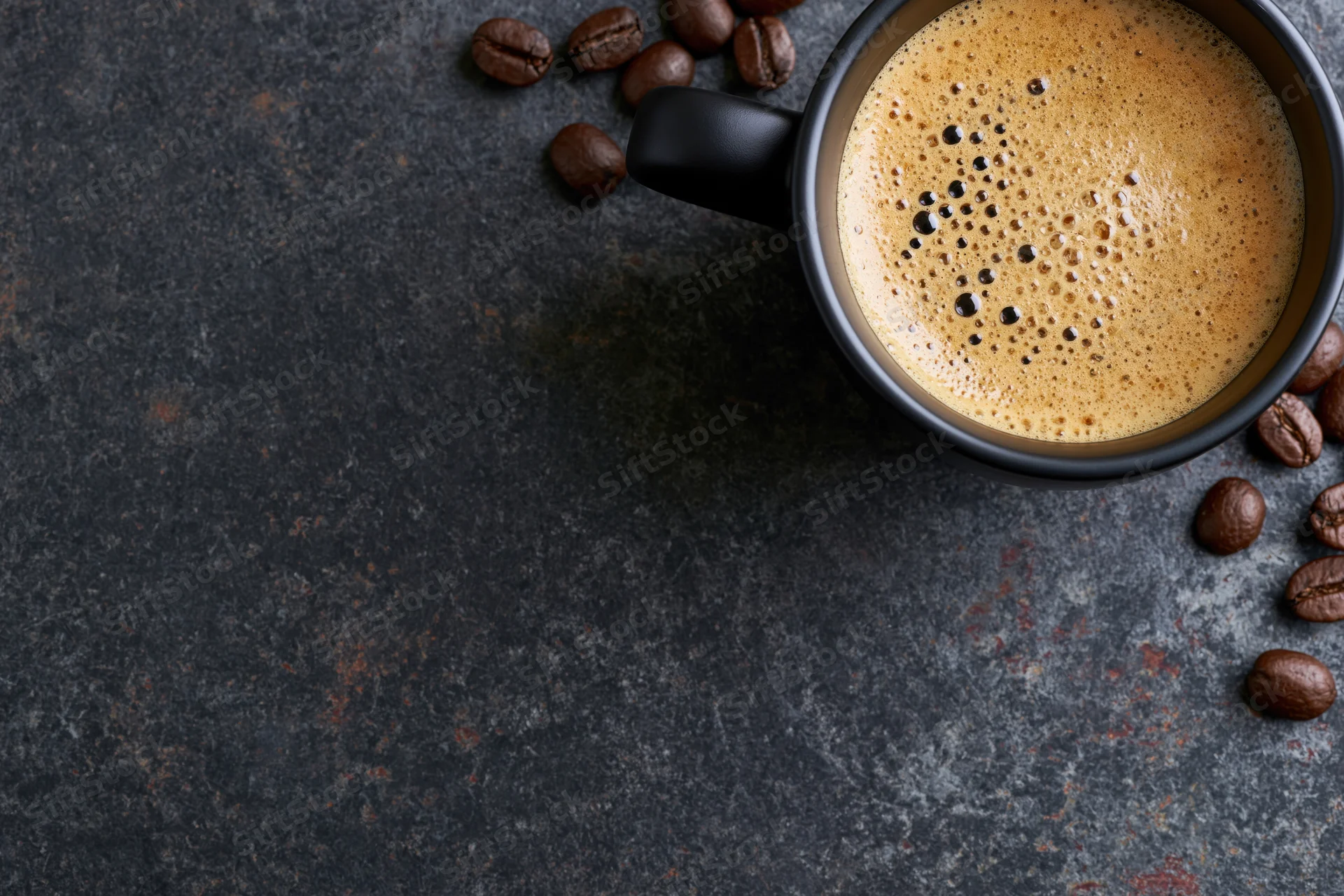 Black ceramic mug filled with frothy espresso coffee on dark textured surface with scattered coffee beans around it
