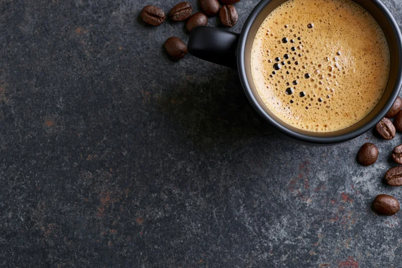 Black ceramic mug filled with frothy espresso coffee on dark textured surface with scattered coffee beans around it