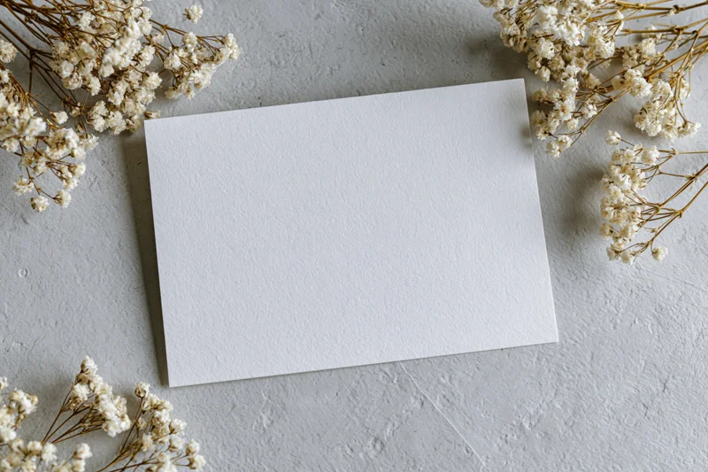 Blank white greeting card lying flat on textured grey surface surrounded by dried baby's-breath flowers