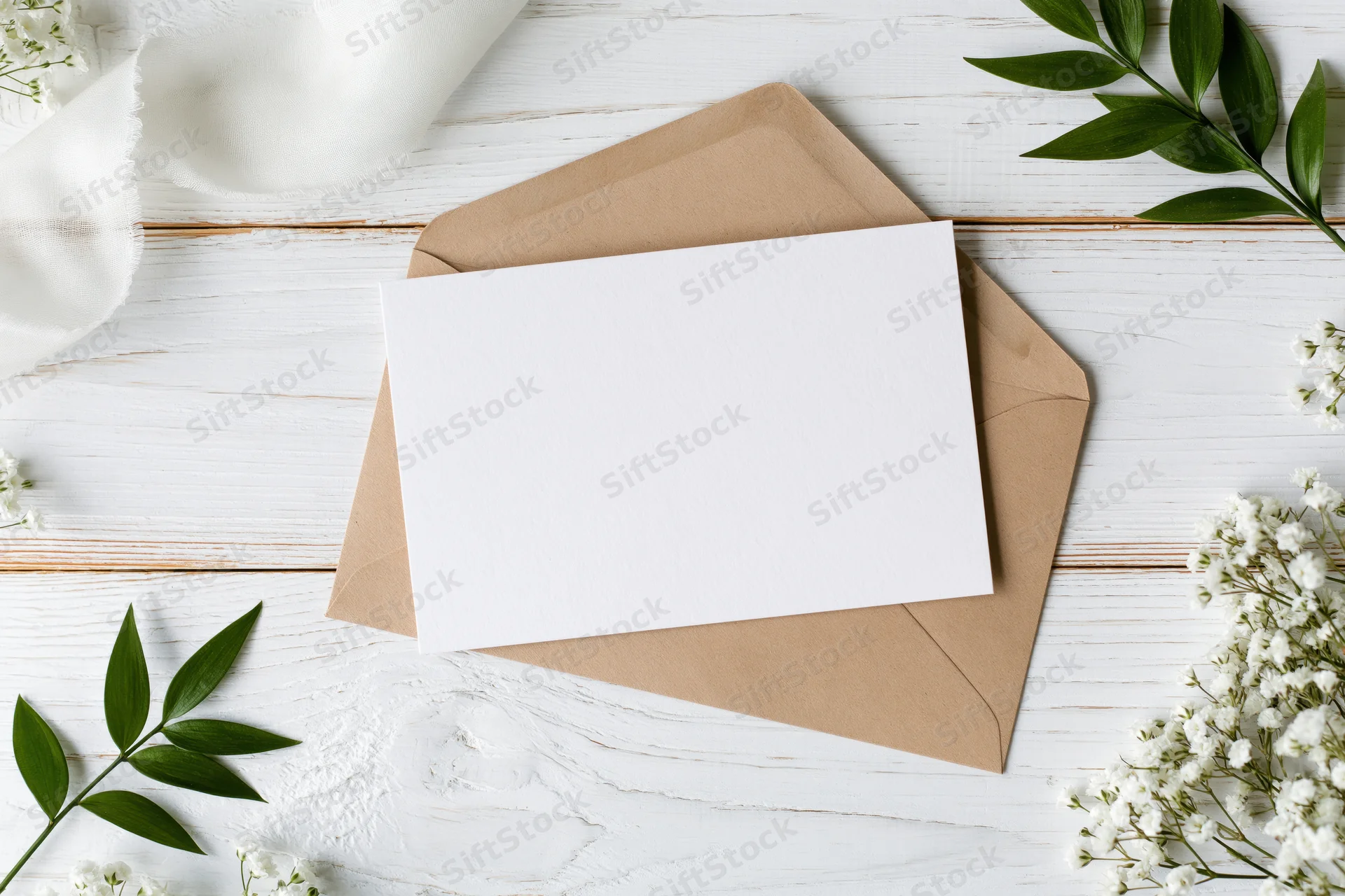 Blank white greeting card resting on a kraft paper envelope on a white wooden surface with green leaves and baby's breath flowers arranged around it