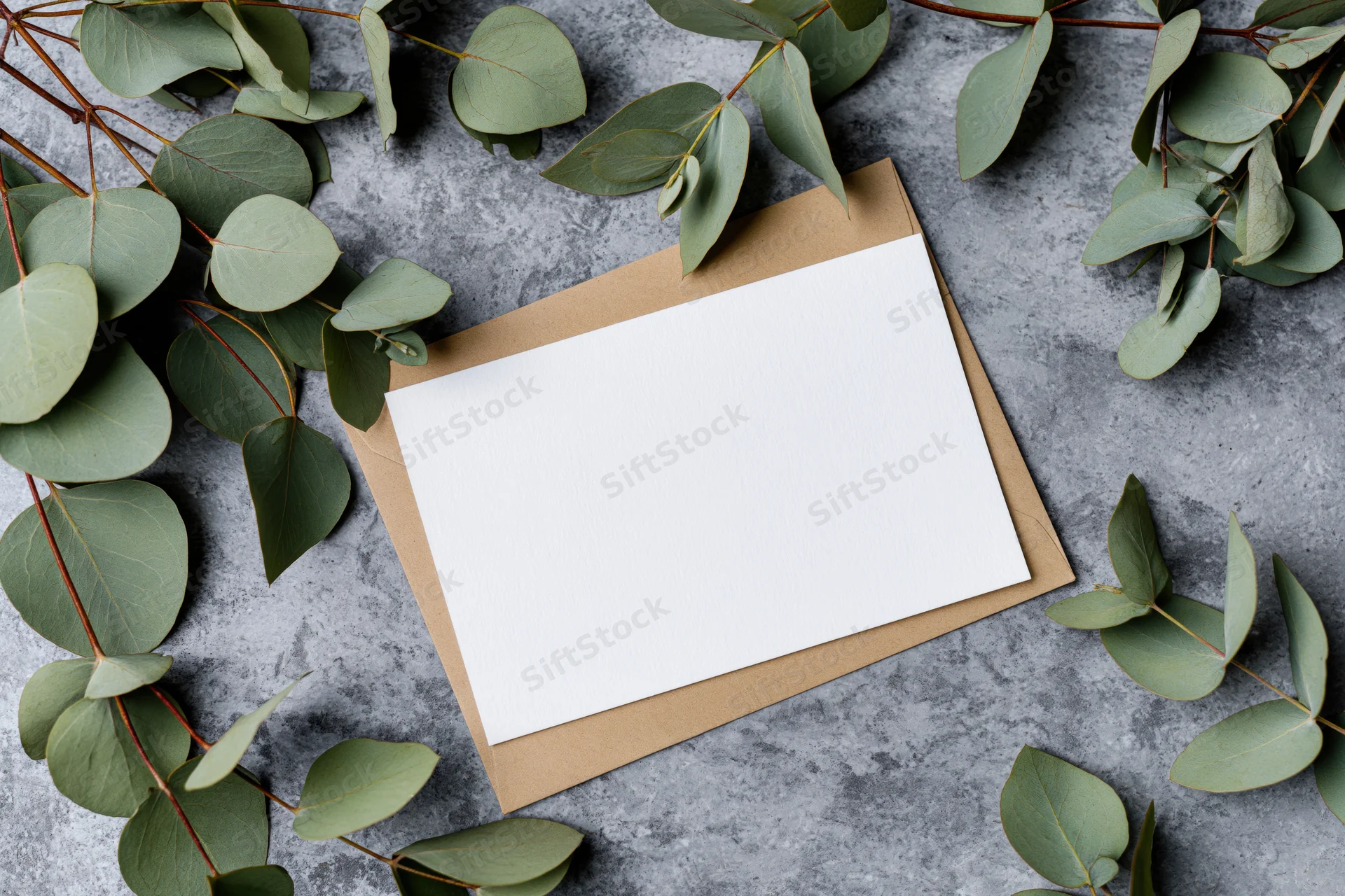 Blank white greeting card resting on kraft envelope surrounded by eucalyptus leaves on gray surface
