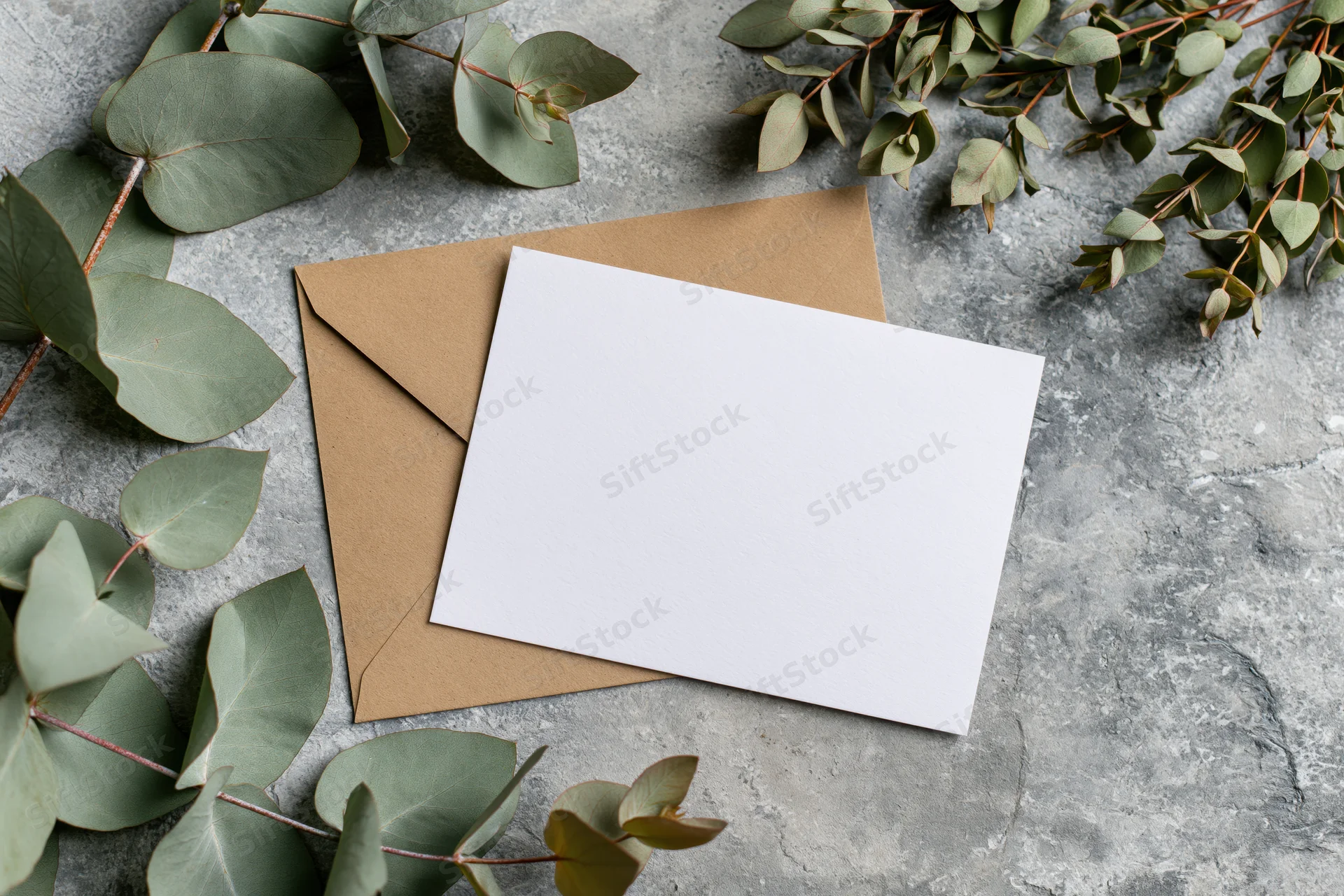 Blank white greeting card resting on kraft envelope with eucalyptus leaves on gray stone surface