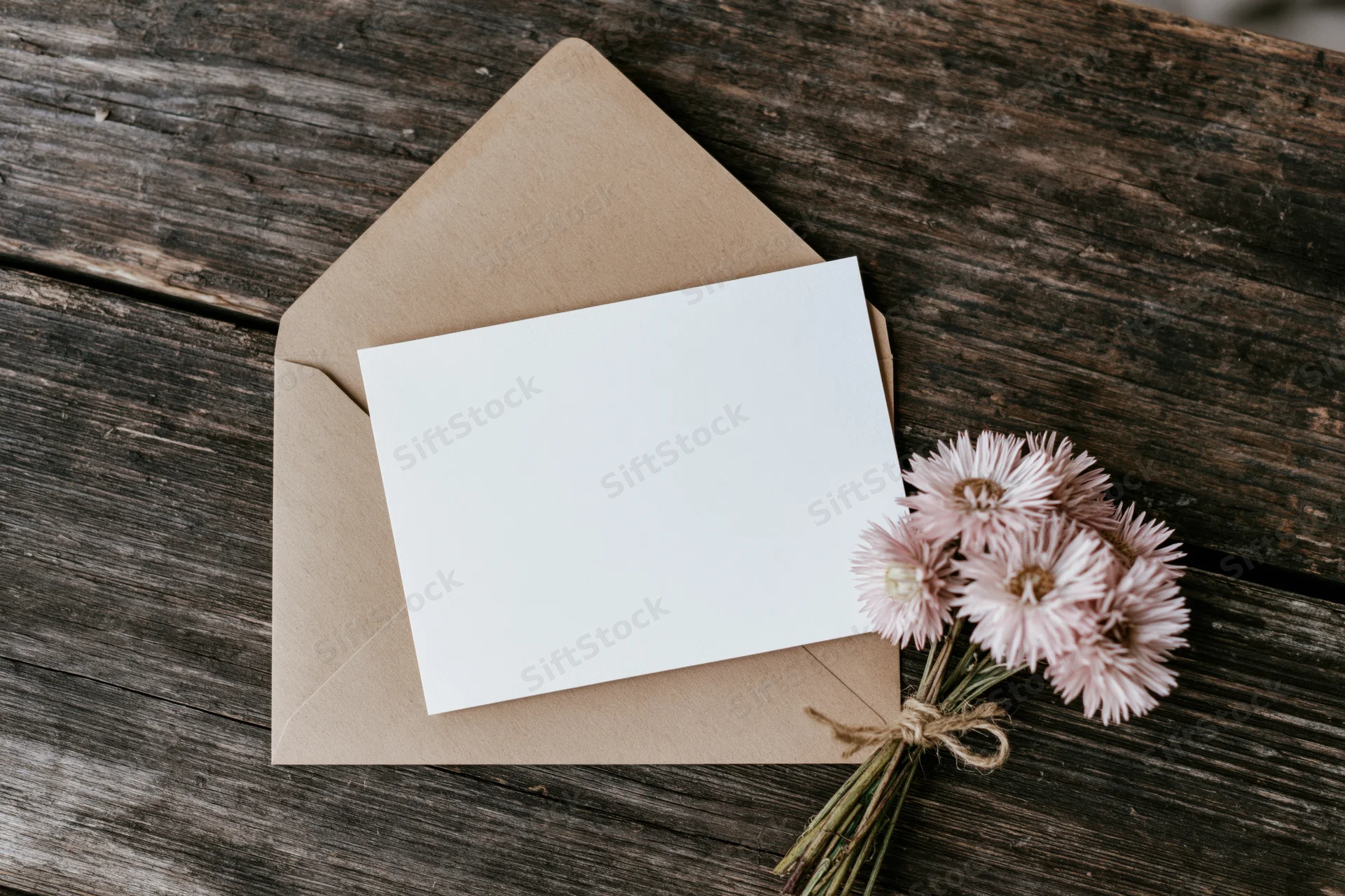 Blank white card resting on open kraft envelope with dried pink flowers tied with twine on wooden surface