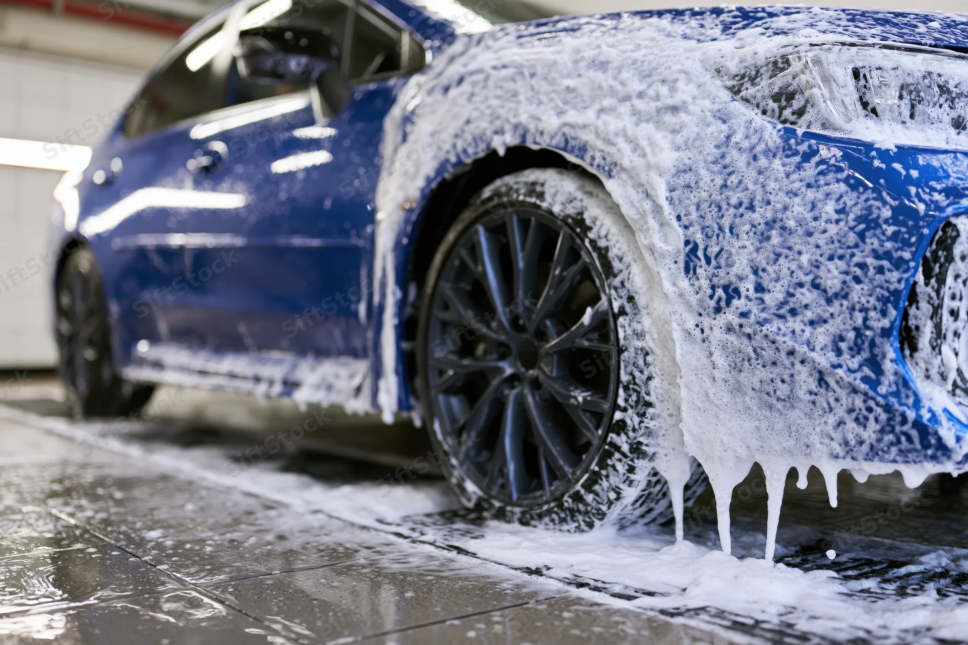 Blue sports car covered in thick white soap foam during a car wash in a garage