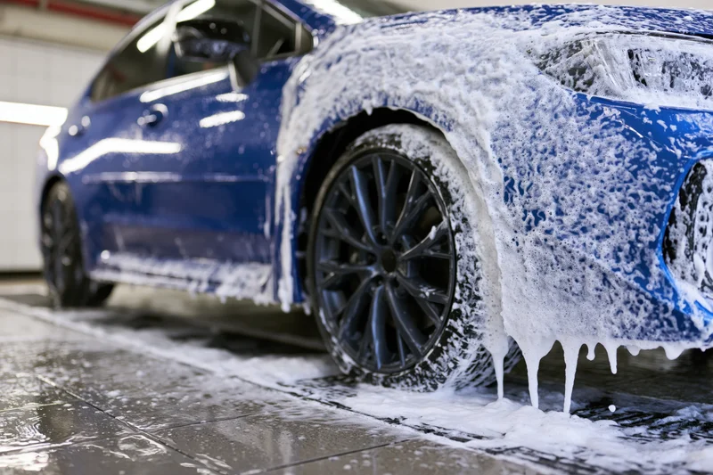 Blue sports car covered in thick white soap foam during a car wash in a garage