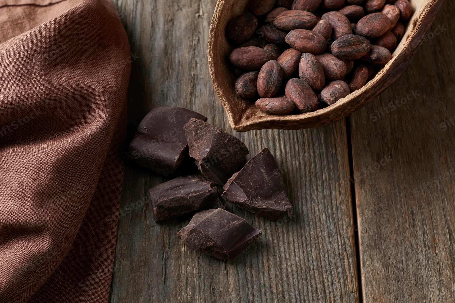 Broken dark chocolate chunks scattered on a weathered wooden surface beside a bowl of cocoa beans and a brown cloth