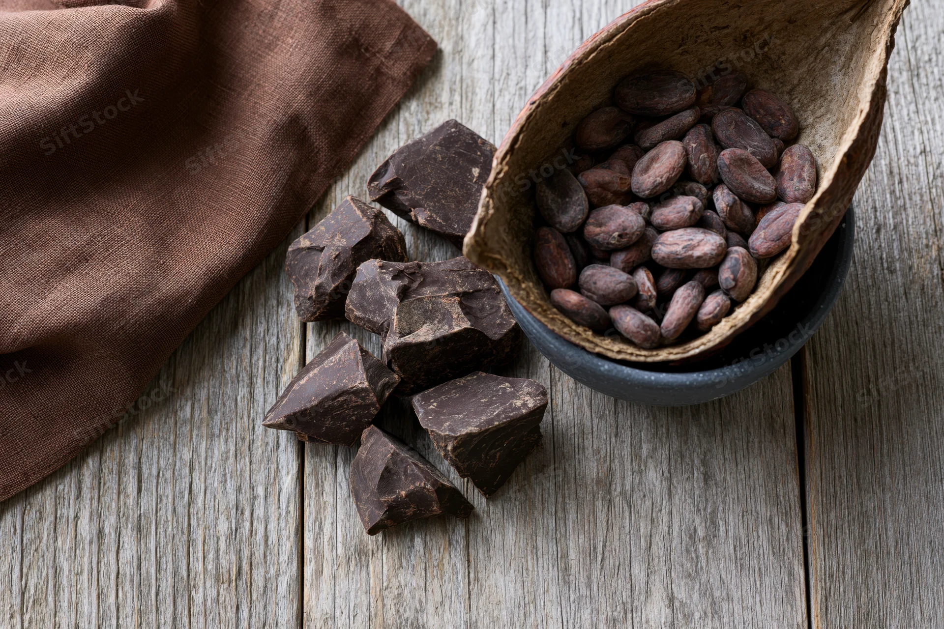 Broken dark chocolate chunks and cocoa beans in a coconut shell bowl on a weathered wooden surface with a brown cloth nearby