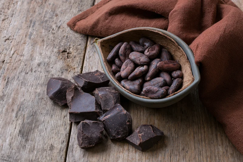 Broken dark chocolate chunks scattered on a weathered wooden surface with cocoa beans in a pod husk beside a brown cloth