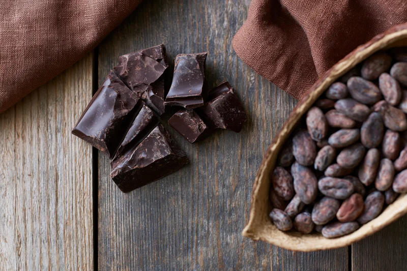 Broken dark chocolate chunks scattered on a weathered wooden surface beside a basket of cocoa beans