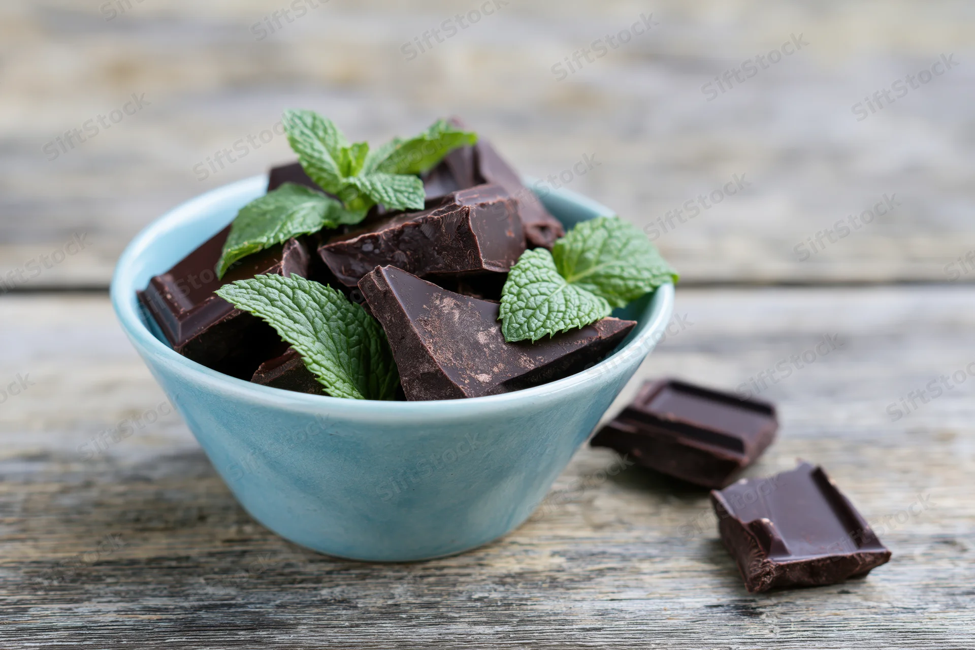 Broken dark chocolate pieces with mint leaves in a blue ceramic bowl on a wooden surface