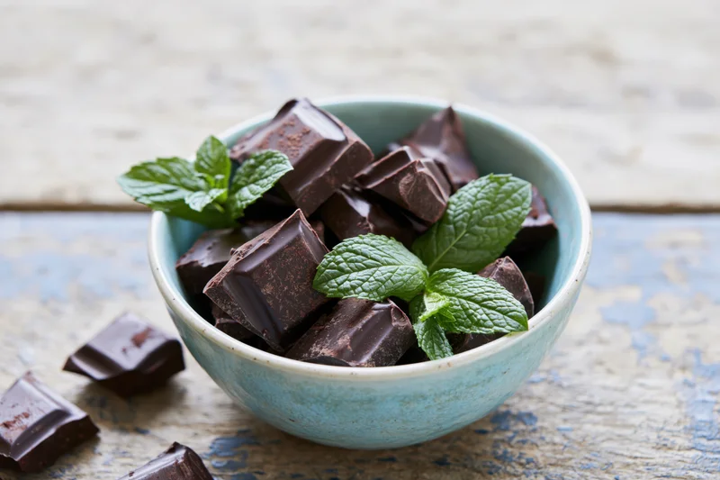 Broken dark chocolate pieces in a small blue ceramic bowl with fresh mint leaves on a rustic wooden surface