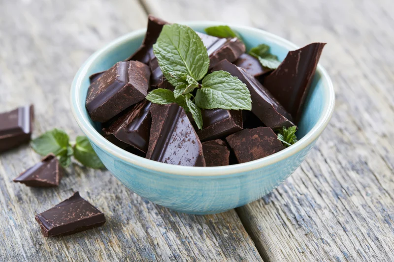 Broken dark chocolate pieces in a small blue ceramic bowl with fresh mint leaves on a rustic wooden surface