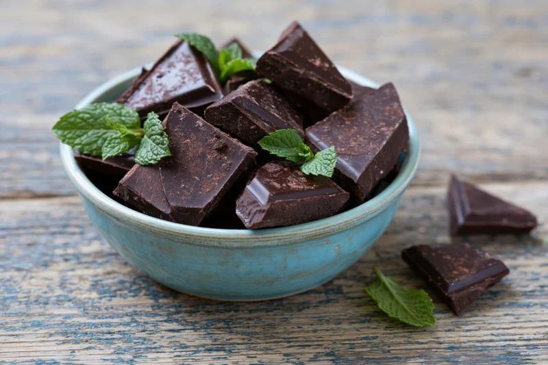 Broken dark chocolate pieces piled in a small blue ceramic bowl with mint leaves on a rustic wooden table