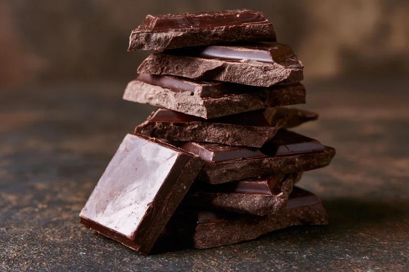 Broken dark chocolate pieces stacked unevenly into a tall tower on a dark textured surface