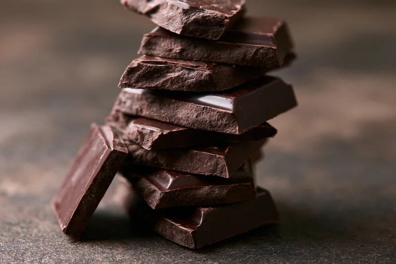 Broken dark chocolate pieces stacked unevenly into a tall tower on a textured surface