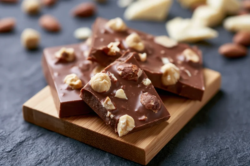 Broken milk chocolate bar pieces with whole hazelnuts arranged on a wooden board with scattered nuts and chocolate chunks on a dark slate surface