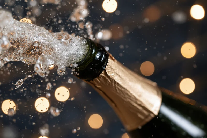 Champagne bottle neck tilted upward mid-pop with liquid droplets and bubbles spraying against bokeh lights