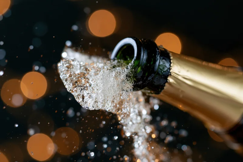 Champagne bottle neck tilted upward mid-pop with liquid droplets and bubbles spraying against dark bokeh background