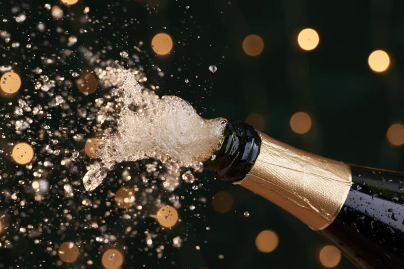 Champagne bottle neck tilted upward mid-pop with liquid droplets and bubbles spraying against a dark bokeh background
