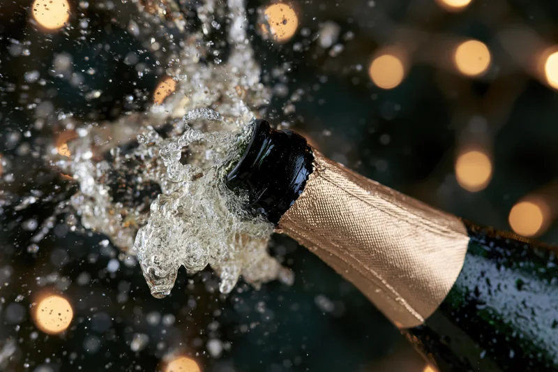 Champagne bottle neck tilted upward mid-pop with liquid droplets and bubbles spraying against blurred bokeh lights