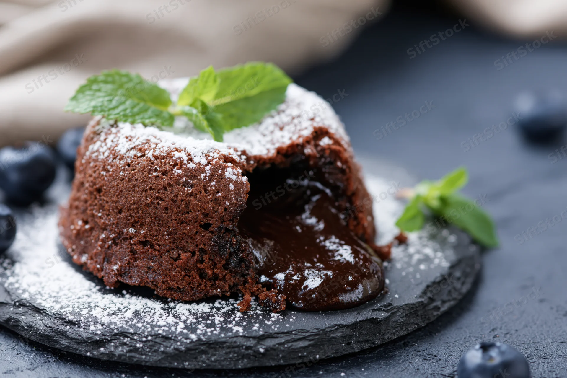 Chocolate lava cake with molten chocolate oozing from a break, garnished with mint and blueberries on a slate plate dusted with powdered sugar