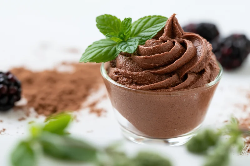Chocolate mousse in a glass cup topped with mint leaves and surrounded by cocoa powder and blackberries on a white surface