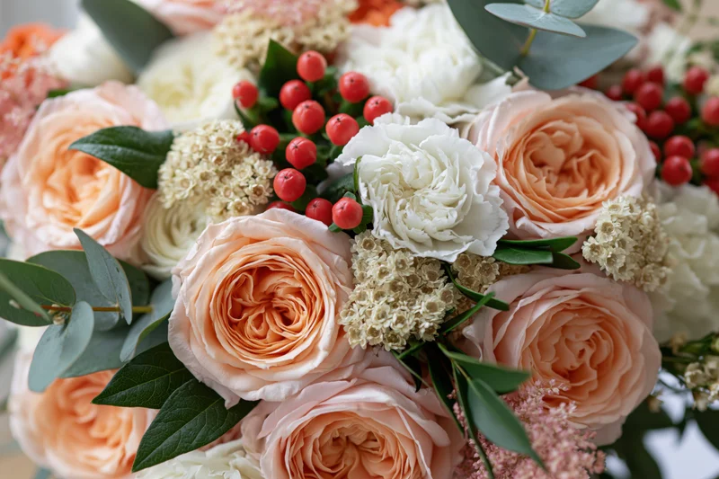 Close-up of a bridal bouquet with peach garden roses, white flowers, greenery and red berries