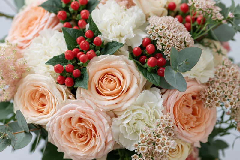Close-up of a bridal bouquet with peach garden roses, white flowers, red berries and green foliage arranged in a soft floral composition