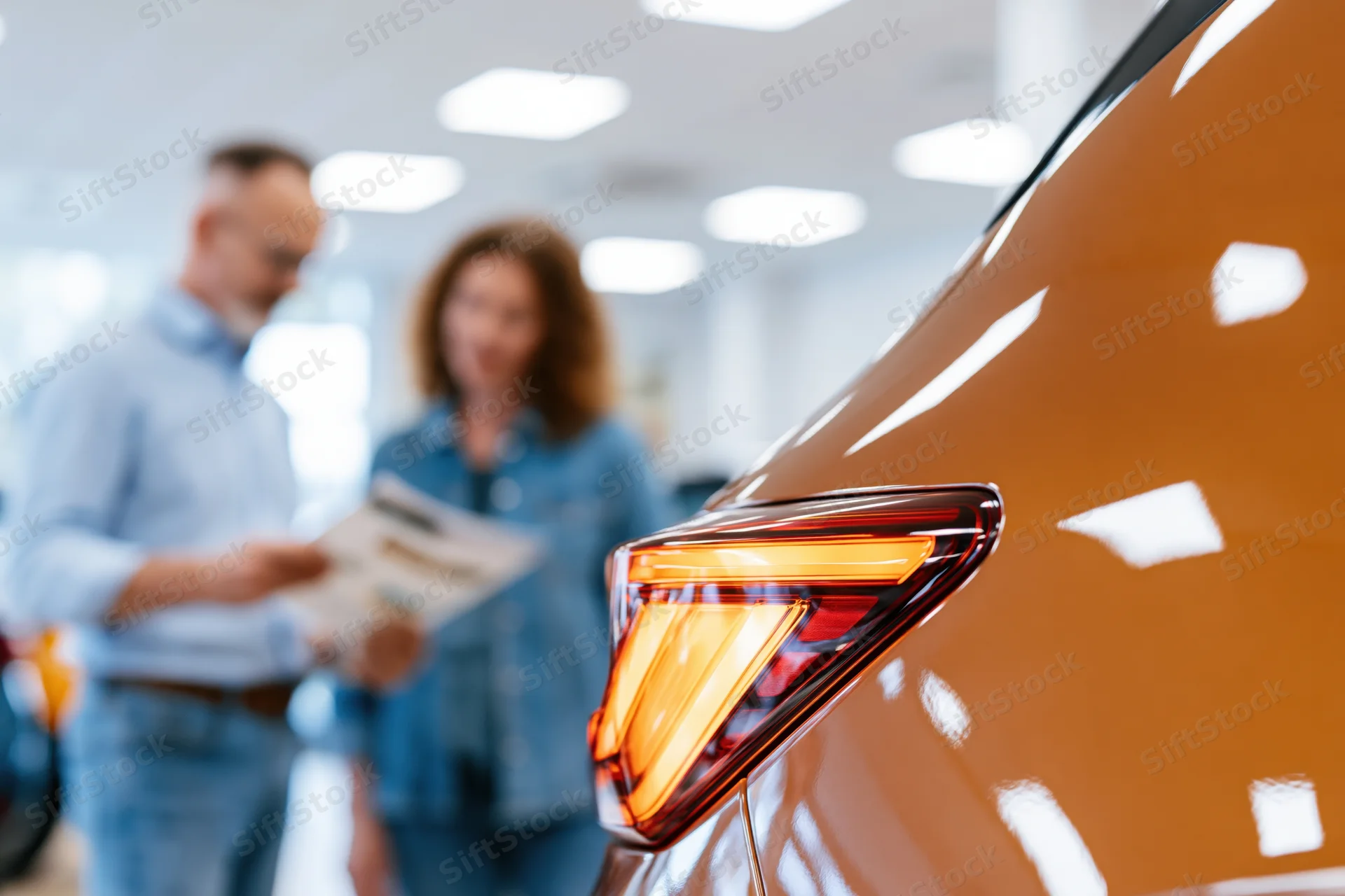 Close-up of copper-brown suv tail light in focus with couple reviewing brochure in showroom background