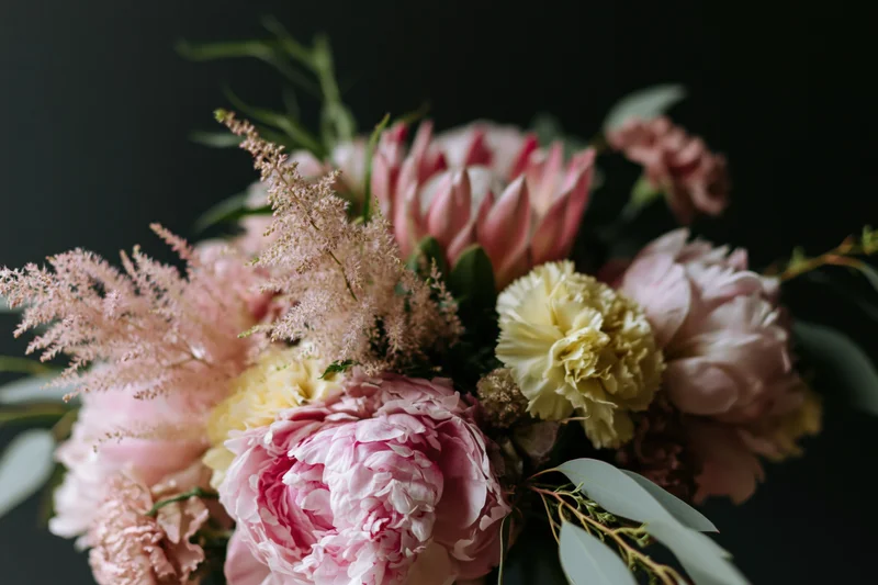 Close-up of a loosely arranged garden bouquet with pink peonies, cream carnations, astilbe and eucalyptus foliage against a dark background