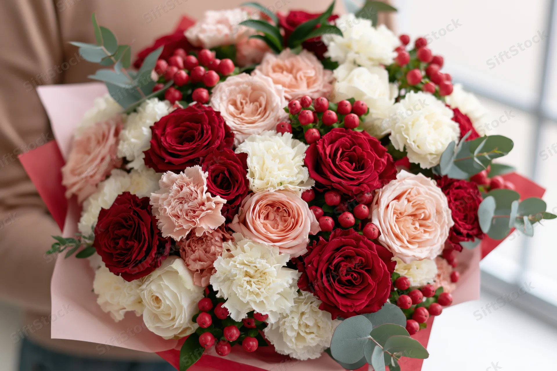 Woman holding a lush mixed bouquet of red roses, white carnations, pink peonies, and eucalyptus with red berries wrapped in pink paper