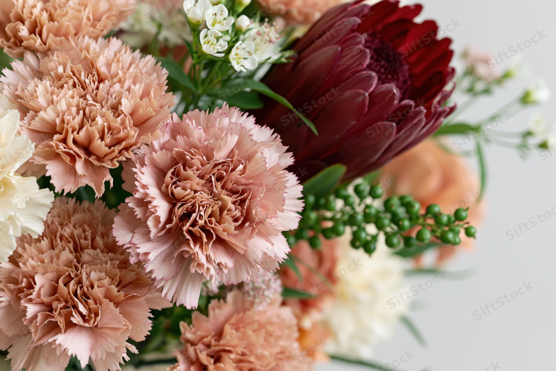 Close-up of a mixed floral arrangement with dusty rose carnations, burgundy dahlia, white filler flowers and greenery on a light background